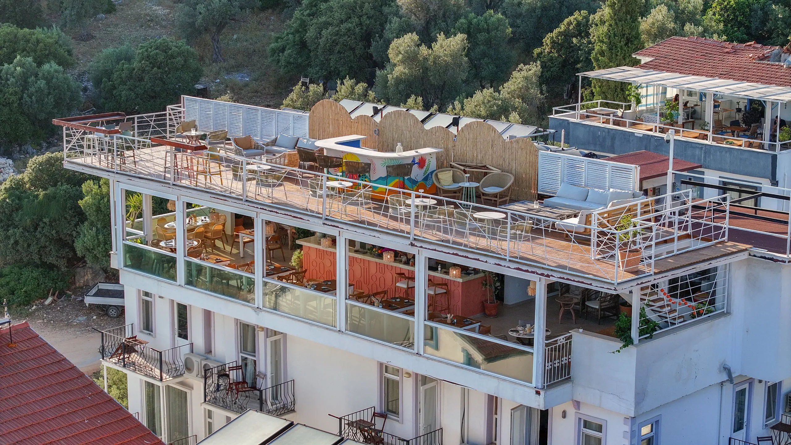 Yukarıdan Koi: beyaz otel binasının çatısında açık teras, altında camlı korunaklı kat ve terakota bar; arkada zeytinlik yamaç.