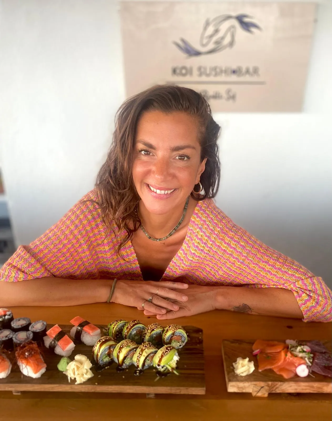Renkli Şef tezgâhın arkasında gülümsüyor; önünde tahta altlıklarda maki, nigiri, avokadolu bir rol ve sashimi, arkasında Koi Sushi Bar tabelası.